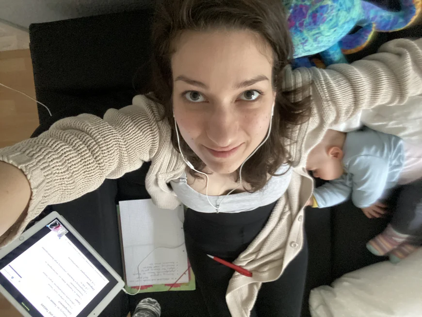 Eine Studentin fotografiert sich aus der Vogelperspektive, sie hat ein Baby im linken Arm, auf der Couch liegen Schreiutensilien und ein Tablet