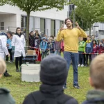 Mann mit gelbem Pullover steht auf einer Wiese und erklärt etwas für eine Gruppe Kinder