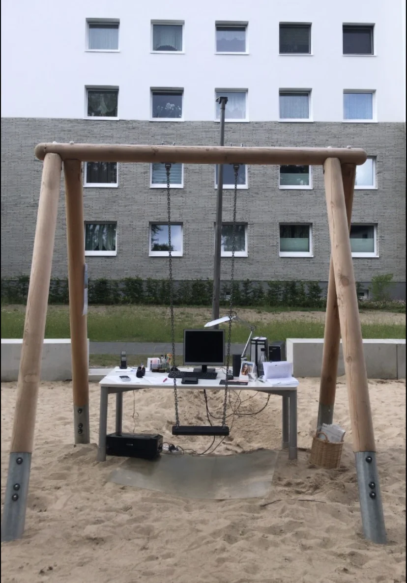 Auf einem Spielplatz unter einer Schaukel im Sand steht ein Schreibtisch, so dass die Schaukel als Stuhl dient. Auf dem SChreibtisch ist ein kompletter Arbeitsplatz eingerichtet, mit Rechner, Schreibtischlampe, Schreibkram. Daneben steht ein Papierkorb, darunter ein Drucker. Hintergrund Plattenbau