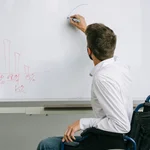 Ein Mann sitzt im Rollstuhl, er trägt ein weißes Hemd und zeichnet etwas auf einem Whiteboard an. 