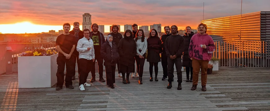 Eine Gruppe junger Menschen steht auf einer Dachterrasse und lächelt in die Kamera. Im Hintergrund sieht man die Dächer Potsdams und einen Sonnenuntergang.