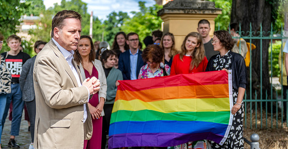 Ein Mann redet, im Hintergrund viele Menschen, es wird eine Regenbogenflagge gezeigt.