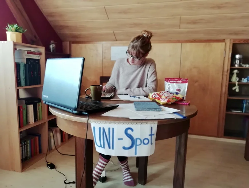 Eine junge Frau sitzt an einem runden Holztisc, darauf steht ein Laptop und es ist ein Schild mit der Aufschrift "Uni Spot" daran befestigt