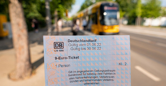 Person hält das 9-Euro-Ticket in der Hand, im Hintergrund ist verschwommen eine gelbe Bahn zu sehen.
