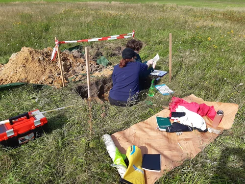 Zwei Personen auf Wiese in gegrabener Grube