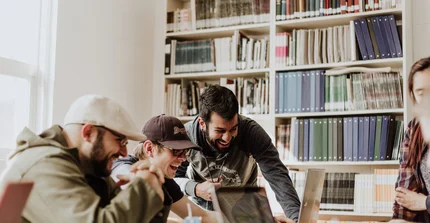 Mehrere Menschen sitzen am Schreibtisch, sie sind sehr gut gelaunt, im Hintergrund sind Bücherregale voller Bücher zu sehen