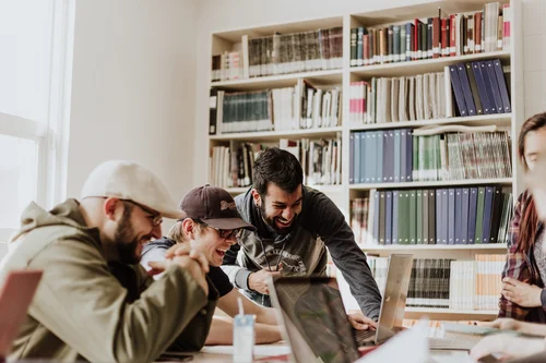 Mehrere Menschen sitzen am Schreibtisch, sie sind sehr gut gelaunt, im Hintergrund sind Bücherregale voller Bücher zu sehen