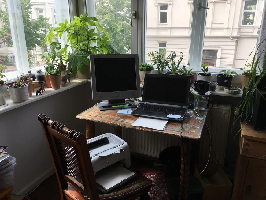 In einer Fensterecke steht ein Schreibtisch. auf der Fensterbank viele Zimmerpflanzen. Der Schreibtisch ist alt und hat Farbflecken. DArauf steht ein alter 4:3 Bildschirm und ein Laptop. Auf dem SChreibtisch stehen Teekanne und Glas. Auf einem Stuhl vor dem Tisch steht ein alter Tintenstrahdrucker-