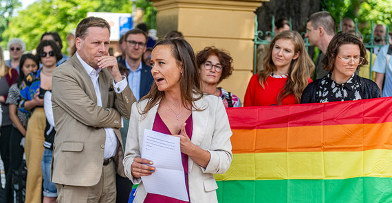 Eine Frau spricht, im Hintergrund etliche Menschen und es wird eine Regenbogenflagge gezeigt.