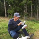 Foto: Scientist sitting on the forest floor, handling the remote control of a white drone standing next to them | Foto: Cosmic Sense consortium