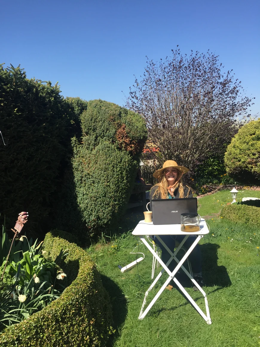 Eine junge Frau sitzt in einem grünen Garten an einem kleinen Tischchen udn arbeitet am Laptop. Sie trägt einen Sonnenhut