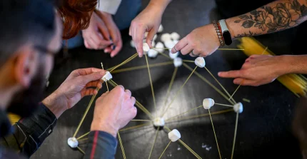 Hände von verschiedenen Personen, die gemiensam aus Marshmallows und Holzstäben etwas bauen.