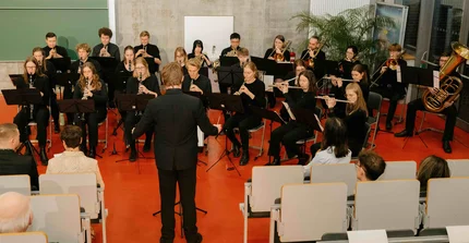 Ein Blasorchester spielt. Alle Personen sitzen. Im Bildvordergrund eine Person, die dirigiert.