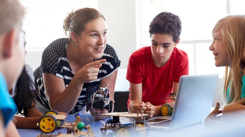 Lehrkraft mit Schüler:innen im Klassenraum. Die Personen stehen um einen Tisch und die Lehrerin zeigt mit dem Zeigefinger lachend auf ein Kind.