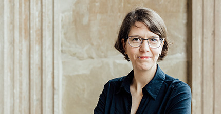 Farbfoto von Gesine Wegner. Weiße Frau in den 30ern mit brauner Kurzhaarfriseur, schwarzer Brille und Lippenpiercing. Sie trägt eine schwarze Bluse und lächelt mit verschränkten Armen in die Kamera. Hintergrund: Torbogen am Neuen Palais.