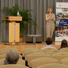 Eine Person steht auf einer Bühne, schräg vor ihr ein Bildschirm, der andere Akteur:innen der veranstaltung und Videos zeigt, im Publikum sitzen vereinzelt Menschen, die Abstand zueinander halten.