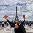 Man sieht glückliche Abolventinnen des Studiengangs mit ihren Diplomen, die sie in die Luft halten, vor dem Eifelturm in Paris.