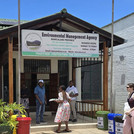 Zu sehen ist das Vordach eines Gebäudes mit drei schwarzen Personen in Bürobekleidung davor und einem großen Schild an der Frontseite, auf dem "Environmental Management Agency - Province Manicaland" steht. Rechts unten im Bild sind drei Mülleimer zur Mülltrennung zu sehen.