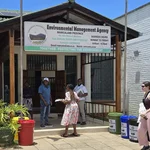 Zu sehen ist das Vordach eines Gebäudes mit drei schwarzen Personen in Bürobekleidung davor und einem großen Schild an der Frontseite, auf dem "Environmental Management Agency - Province Manicaland" steht. Rechts unten im Bild sind drei Mülleimer zur Mülltrennung zu sehen.