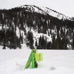 Field site Esterberg: A bright green CRNS sensor with solar panel standing on a white plain with black and white forested mountains in the background | Photo: Cosmic Sense Consortium