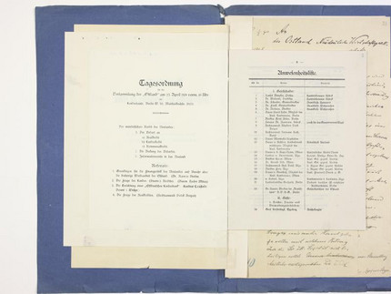 Ein Scan einer Doppelseite. Auf der linken Seite befindet sich ein Zettel überschrieben mit "Tagesordnung" auf der rechten Seite eine Anwesenheitsliste. Alles ist in Sütterlinschrift verfasst.
