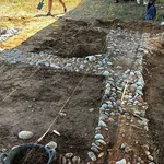 Bild einer Ausgrabungsstelle mit Steinfragmenten, die den Grundriss eines Hauses bilden.