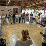 Auf dem Foto zu sehen sind schwarze und weiße junge Erwachsene in einem überdachten Pavillon, die in einem Kreis stehen und Schnüre in den Händen halten, die sich zwischen ihnen als großes Netz aufspannen.