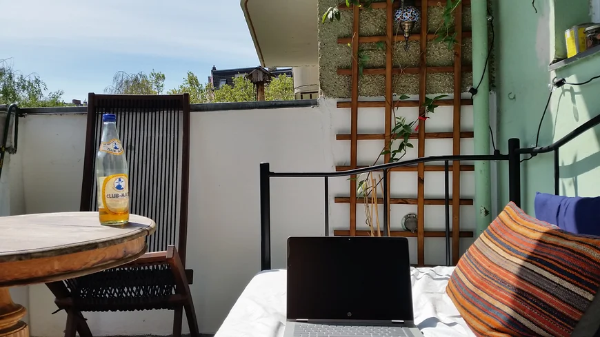Auf einem balkon steht ein Laptio auf einem Sofa, daneben ein Tisch, ein paqar andere balkonmöbel. Es ist sonnig