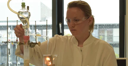 Studierende mit Schutzbrille und Laborkittel in der Hand einen Glaskolben