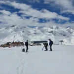 Field site Hohe Mut: Three scientists with snow shoes standing on a flat summit, digging a snow pit. In the background is the top station of a ski lift and a mountain crest | Photo: Cosmic Sense Consortium