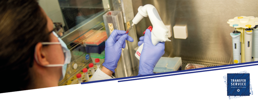 Zwei Hände in blauen Handschuhen mit Pipette in ein Laborgefäß tröpfeln hinter Glasscheibe