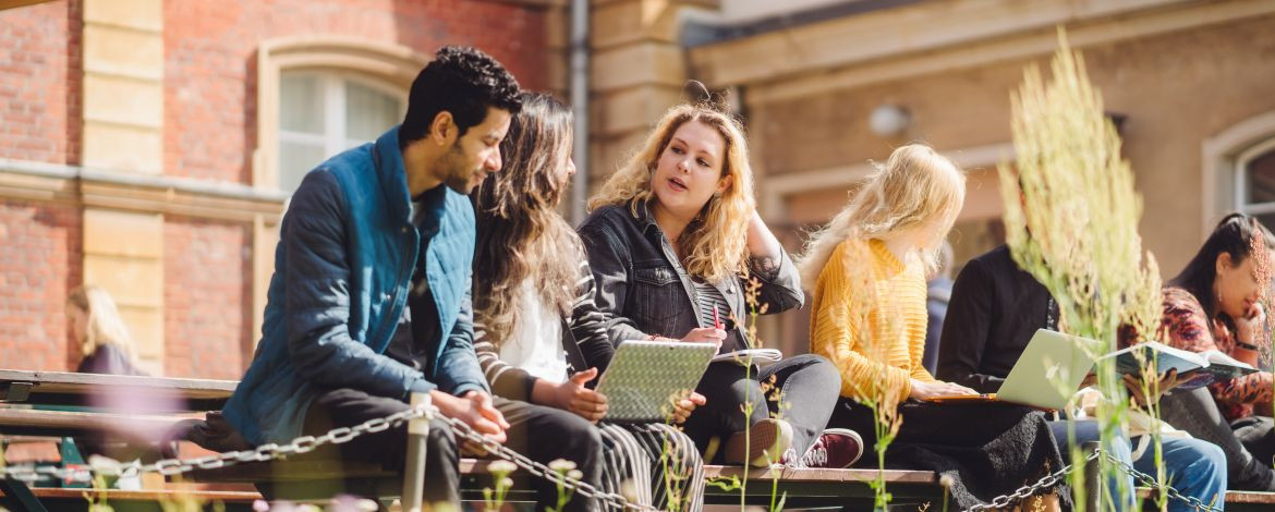 Studierende sitzen auf einer Bank am Campus Neues Palais und unterhalten sich. Eine Studentin hat einen Laptop auf dem Schoß. - 