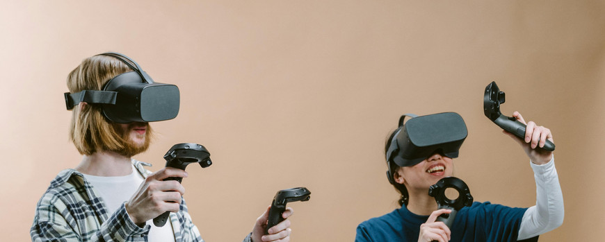 Farbfoto zweier Personen die VR-Brillen tragen. Sie halten jeweils zwei Controller in den Händen und strecken diese nach oben.