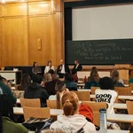 Auf dem Bild sind die Teilnehmenden auf der Bühne im Gespräch zu sehen, während die Studierenden im Publikum aufmerksam zuhören.u.
