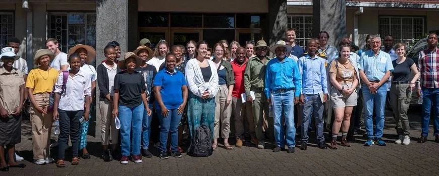 Zu sehen ist eine Gruppe von ca. 25 Personen, die vor einem Gebäude steht. Die Gruppe besteht aus Menschen unterschiedlichen Geschlechts und mit Schwarzer und weißer Hautfarbe. Die meisten Personen blicken lächelnd in die Kamera. Sie tragen legere Kleidung und teilweise Safarihüte gegen die prall scheinende Sonne.