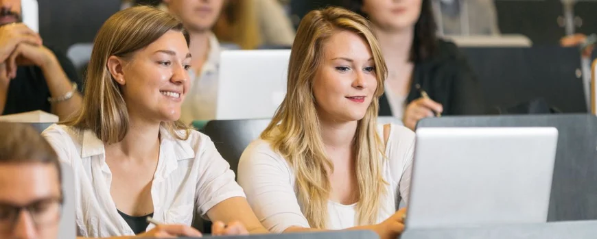 Zwei Studentinnen sitzen mit einem Laptop in einem Hörsaal und lächeln. Um sie herum sitzen weitere Studierende.