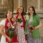 Drei Absolvent*innen posieren mit Blumensträßen vor den Säulen des Kolonnadenbogens.