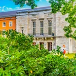 View of the portal of a large house framed by bushes, in front of which is a figure of Little Red Riding Hood