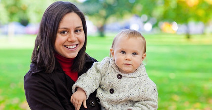 Eine junge Frau mit einem kleinen Kind auf dem Arm. Im Hintergrund eine große Grünfläche.