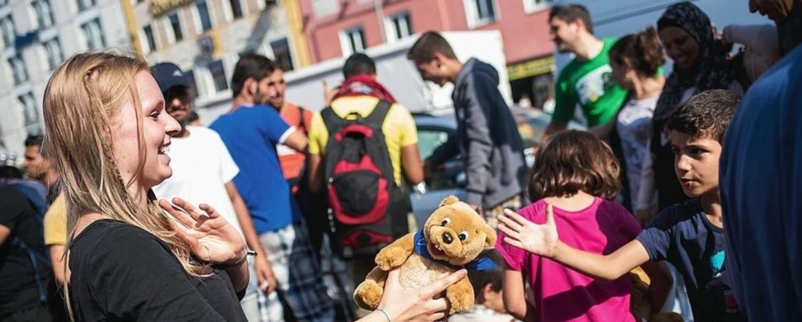 Es sind mehrere Menschen abgebildet. Darunter eine junge Frau, die einem ausländischen Jungen ein Kuscheltier gibt. - 