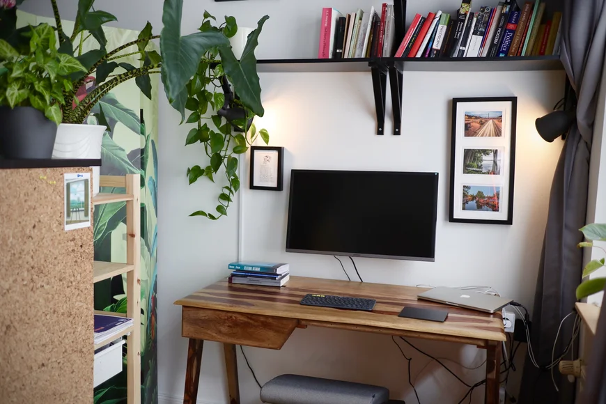 Aufgeräumter Schreibtisch mit Bildschirm an der Wand dahinter befestigt. Links im Anschnitt ein Regal auf dem viele Zimmerpflanzen stehen, deren Blätter und Äste in den Raum hinein ragen. Auf dem Tisch Laptop und Bücherstapel. Über dem Schreibtisch BÜcherregal mit Fachliteratur