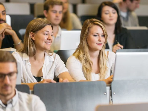 Zwei Studentinnen sitzen mit einem Laptop in einem Hörsaal und lächeln. Um sie herum sitzen weitere Studierende.