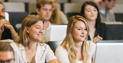 Two female students are sitting in a lecture hall with a laptop and smiling. Seated around them are other students.