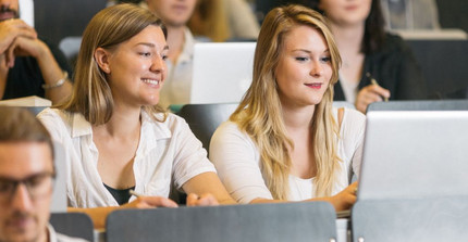 Zwei Studentinnen sitzen mit einem Laptop in einem Hörsaal und lächeln. Um sie herum sitzen weitere Studierende.