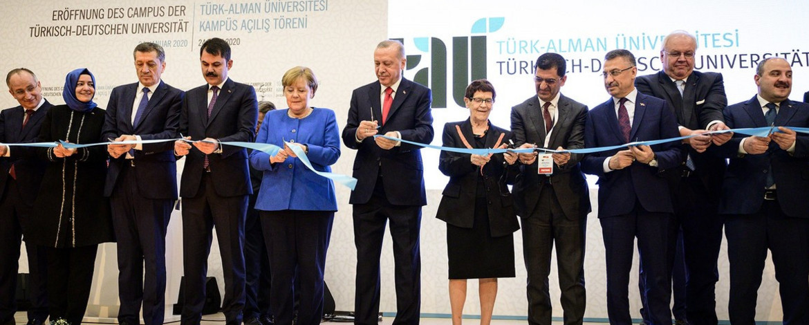 Auf dem Foto ist unter Anderem ehemalige Bundeskanzlerin Frau Dr. Angella Merkel sowie der Staatspräsident der Türkei Recep Tayyip Erdoğan zu sehen. Es wird auf der Bühne ein blaues Eröffnungsband durchgeschnitten. Im Hintergrund sind deutsche und türkische Flagge sowie das Logo der Türkisch Deutschen Universität zu sehen. - 