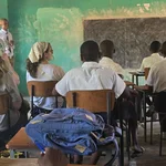 Auf dem Foto sind einige Personen in einem alten Schulraum mit Kreidetafel von hinten zu erkennen. Sie sitzen auf Holzstühlen und schauen nach vorn zur Tafel. Die simbabwischen Schüler:innen tragen weiße Hemden, die deutschen Studentinnen tragen sommerliche Kleidung. Im Hintergrund steht ein Lehrer mit Hemd und Krawatte.