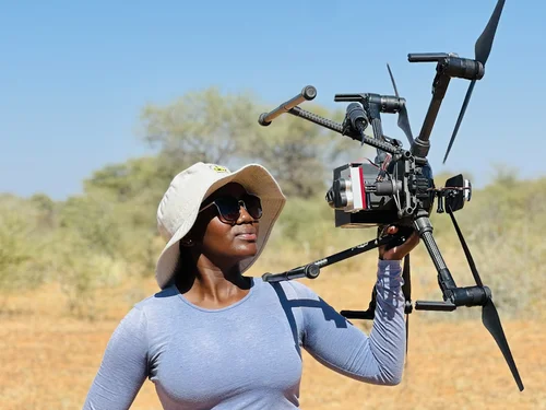 PhD student Vistorina Amputu holding a drone in her hand, with savanna rangeland in the background