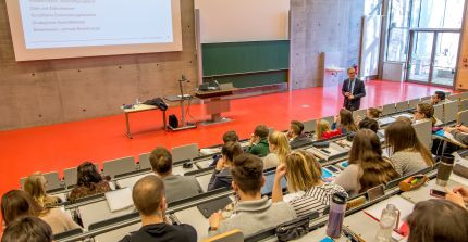 Ein Mann steht in einem Hörsaal vor vielen Studierenden und hält eine Vorlesung