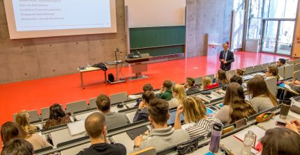 Ein Mann steht in einem Hörsaal vor vielen Studierenden und hält eine Vorlesung