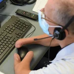 Ein Mann trägt eine Maske, an seinen Händen erkennt man, dass nicht alle Finger ausgebildet sind. Er arbeitet am Computer mithilfe einer speziellen Maus.